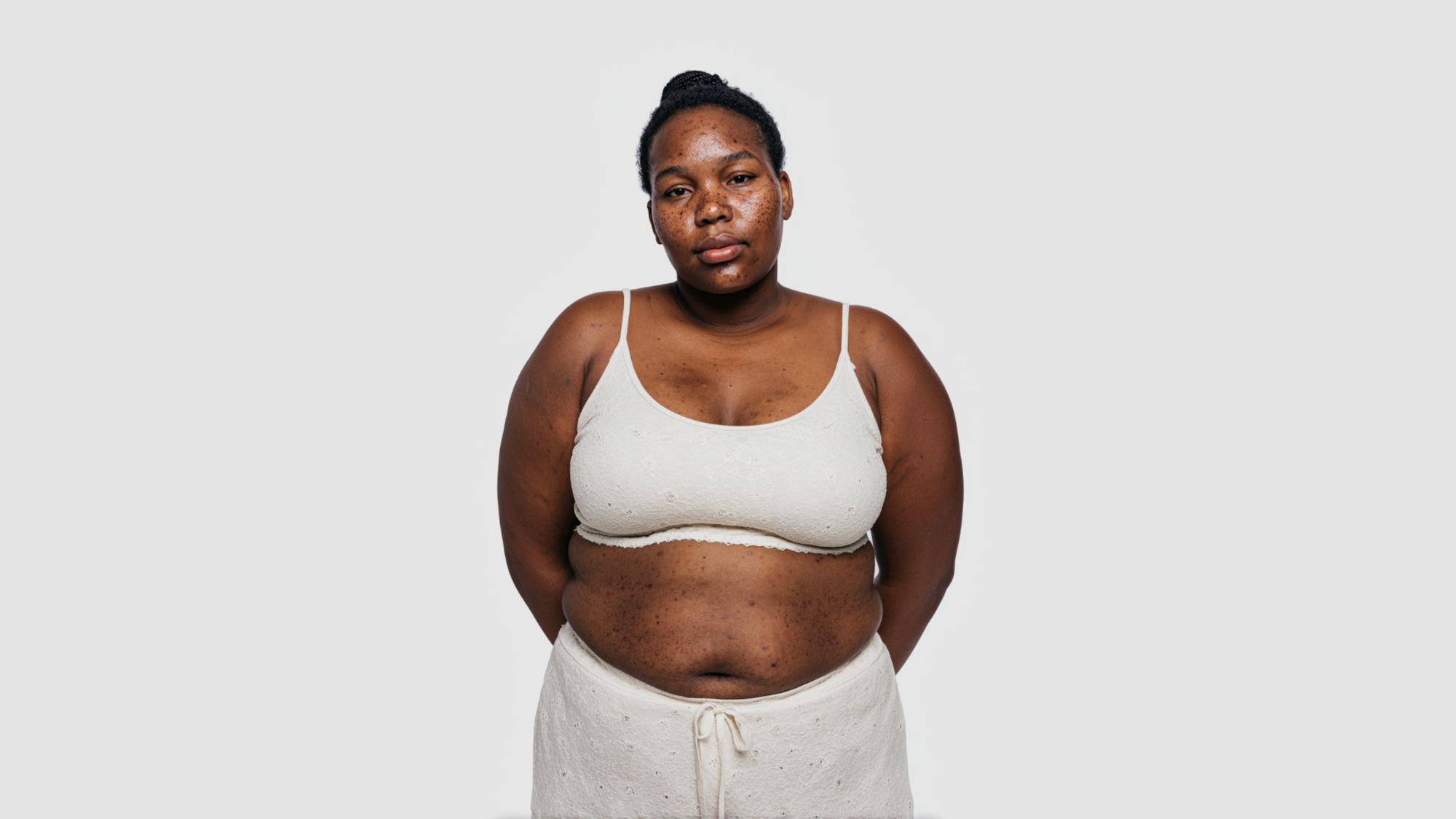 AI-generated full-length view of a fictional woman standing calmly in cream underwear against a grey studio backdrop