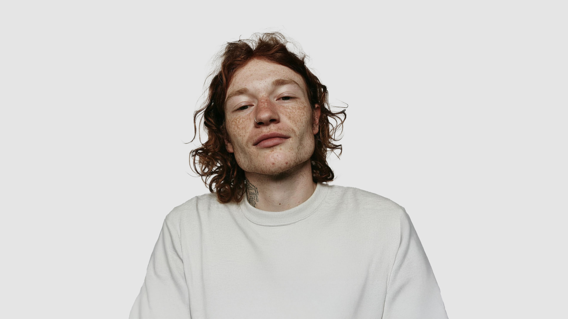 AI-generated front view of a fictional man with long red curly hair, heavy freckles, a nose ring and a small neck tattoo, wearing an off-white sweatshirt against a pale grey background