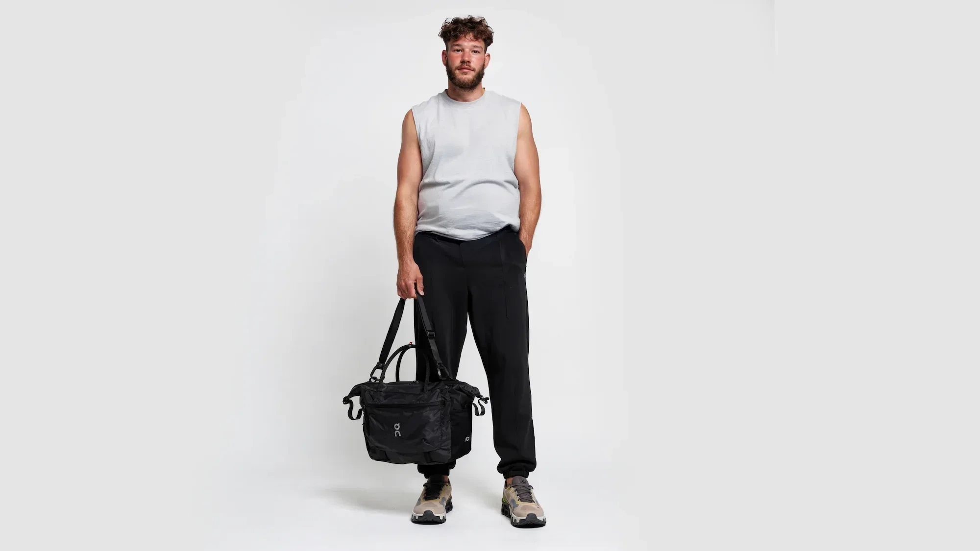 AI-generated full-length view of the same fictional man in a grey sleeveless top and black trousers, holding a black holdall