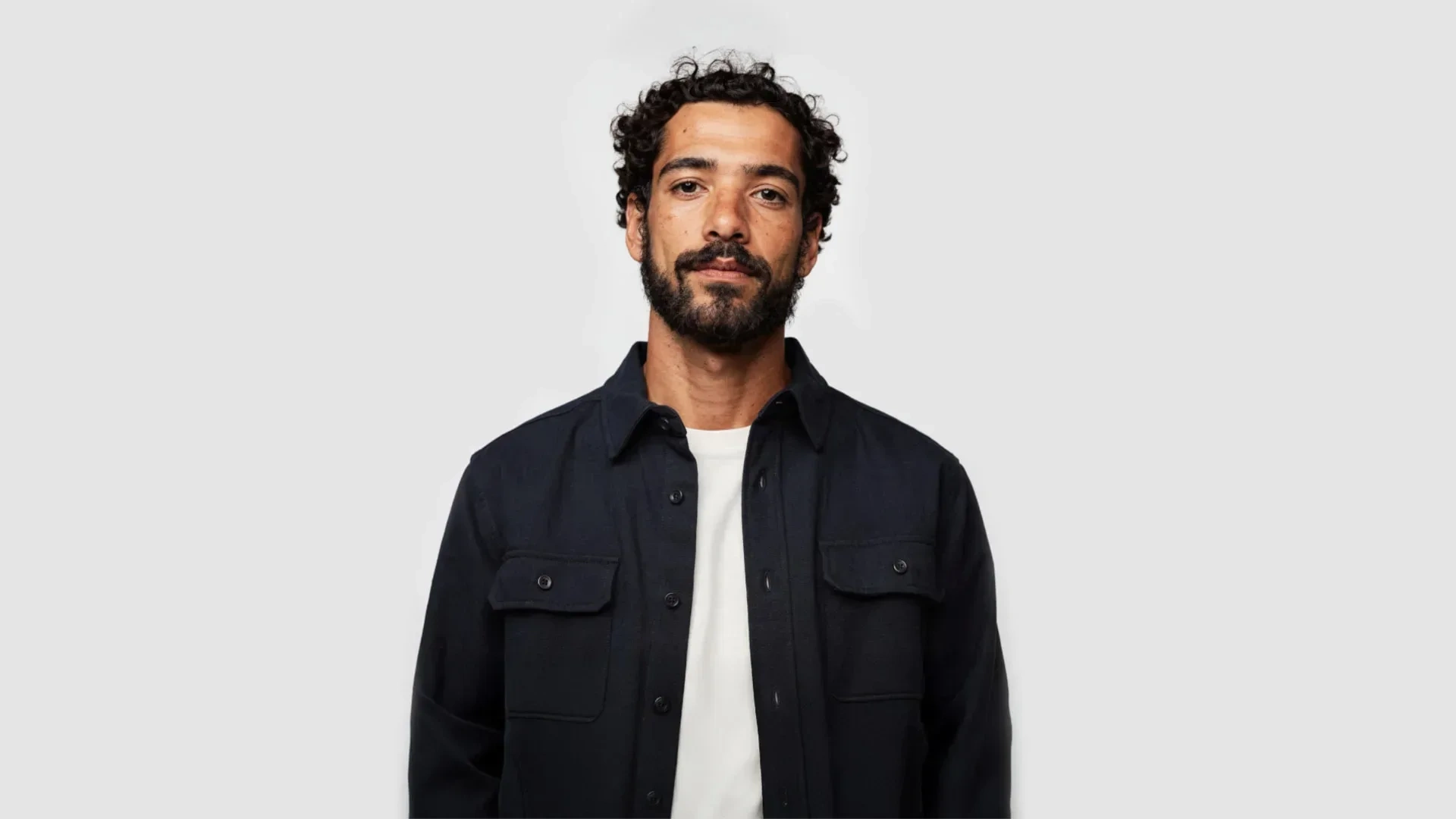 AI-generated front view of a fictional man with dark curly hair and a beard, wearing a navy overshirt over a white t-shirt against a pale grey background