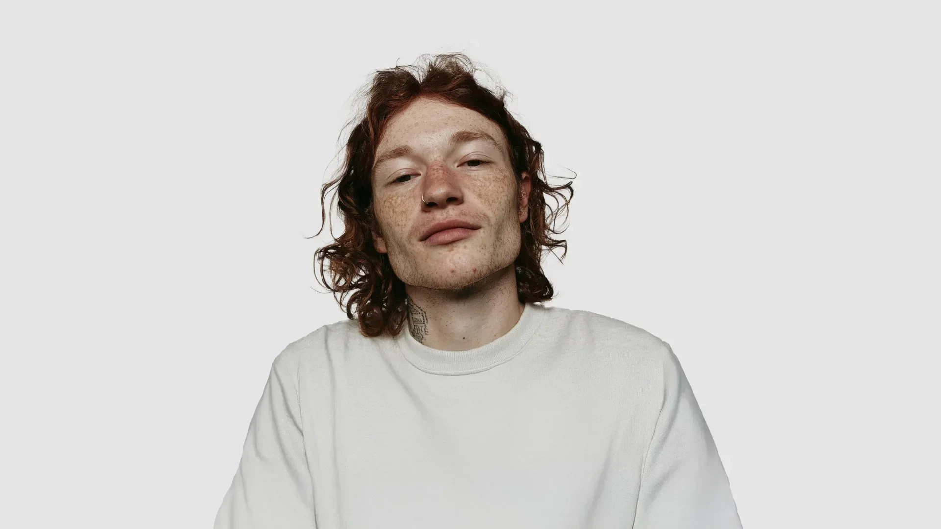 AI-generated front view of a fictional man with long red curly hair, heavy freckles, a nose ring and a small neck tattoo, wearing an off-white sweatshirt against a pale grey background