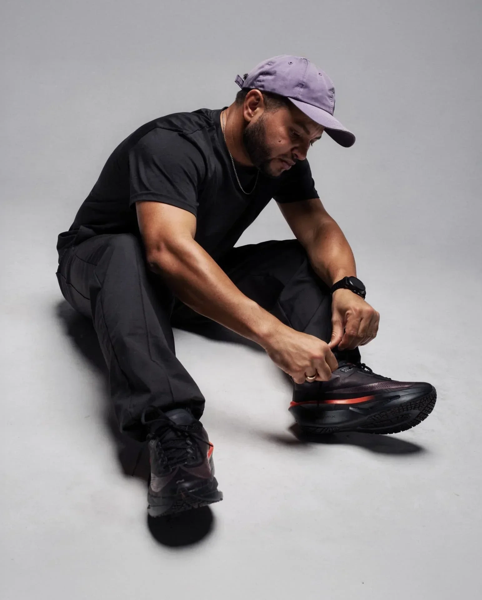 AI-generated view of the same fictional man sitting on a grey studio floor in black clothing, tying the laces of a running shoe