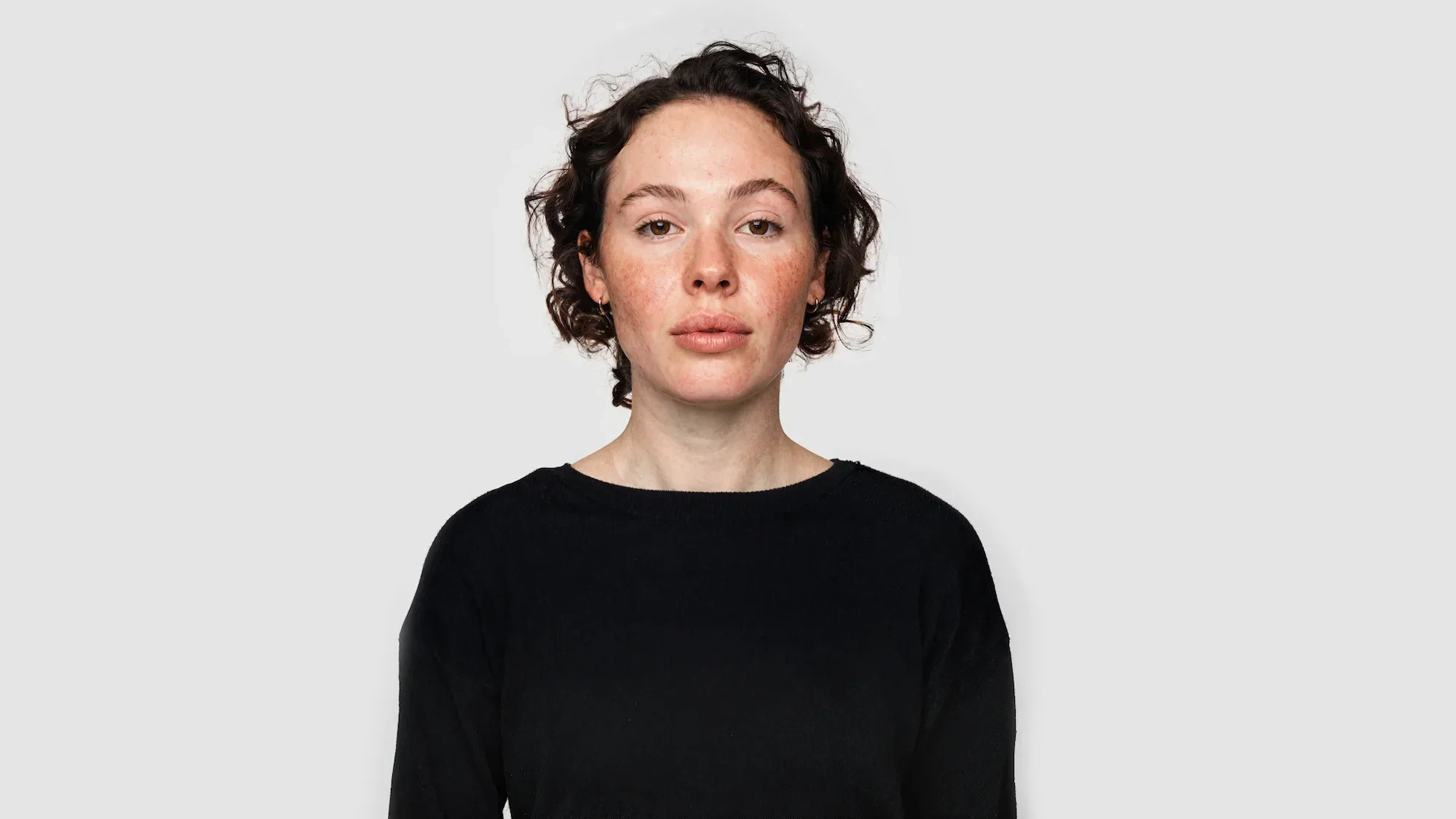 AI-generated front view of a character with freckled skin and short curly dark hair, wearing a black knit against a light grey studio background