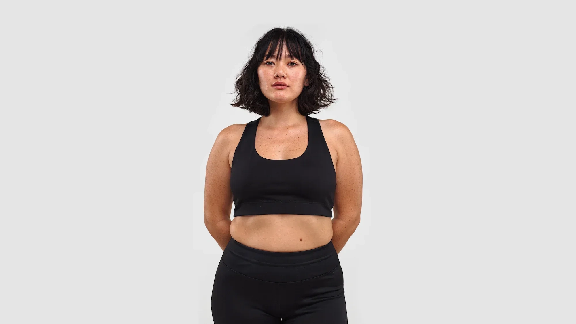 AI-generated front view of a character with short dark wavy hair and freckled skin, standing in plain black sportswear against a light grey studio background