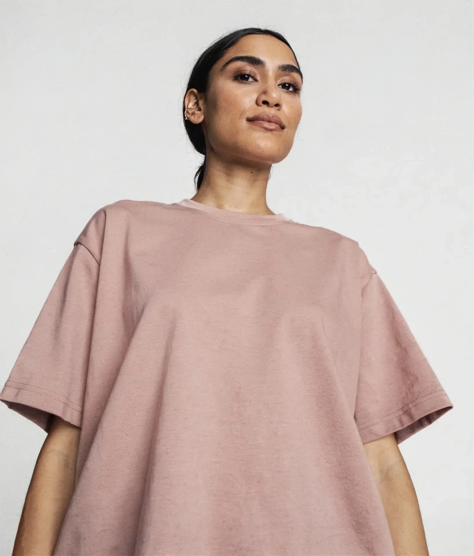 AI-generated portrait of a fictional woman with dark hair tied back, wearing an oversized dusty-pink t-shirt against a pale studio background