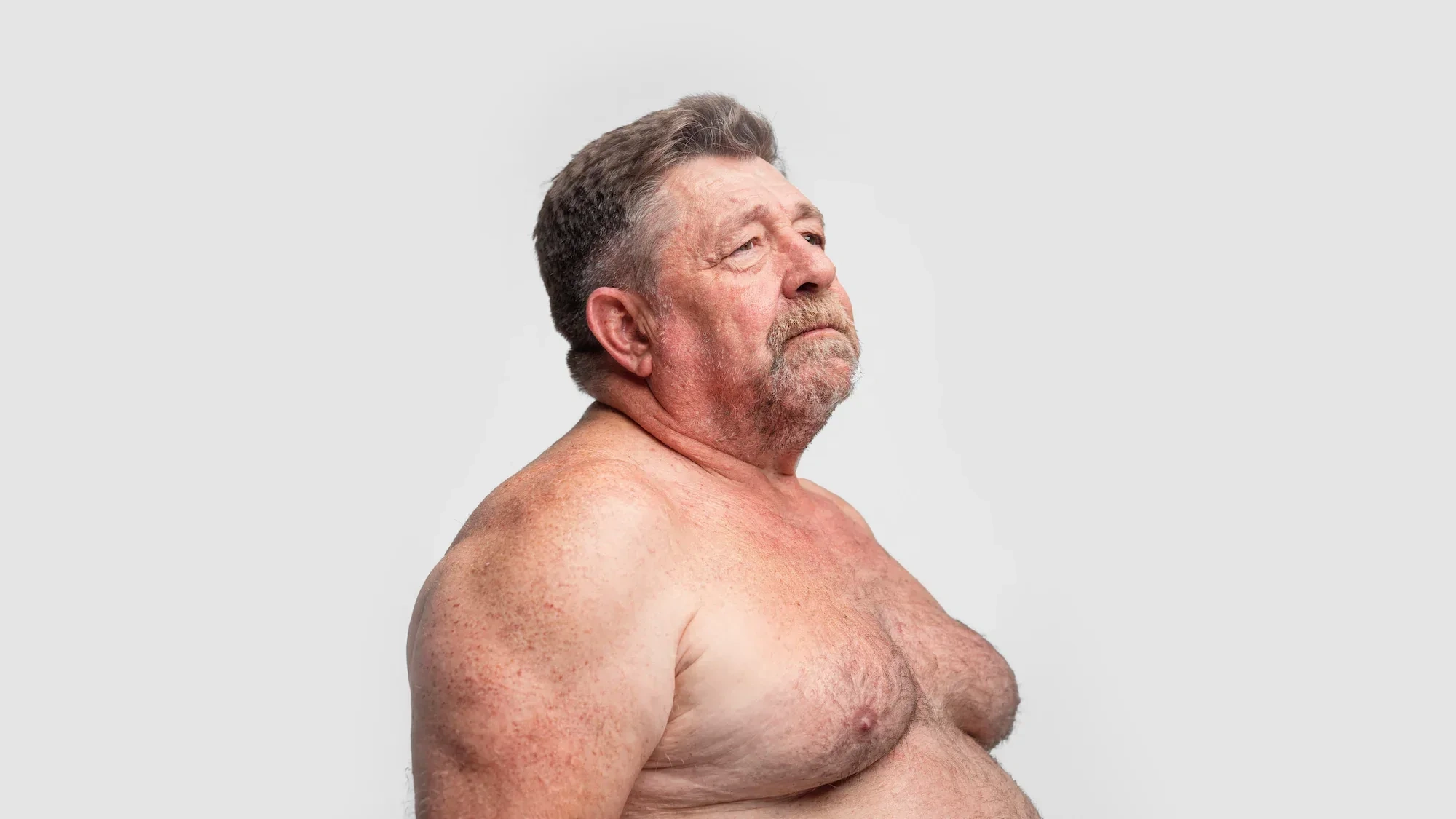 AI-generated three-quarter view of a fictional older man with a grey moustache, bare chest and soft build, against a light studio background